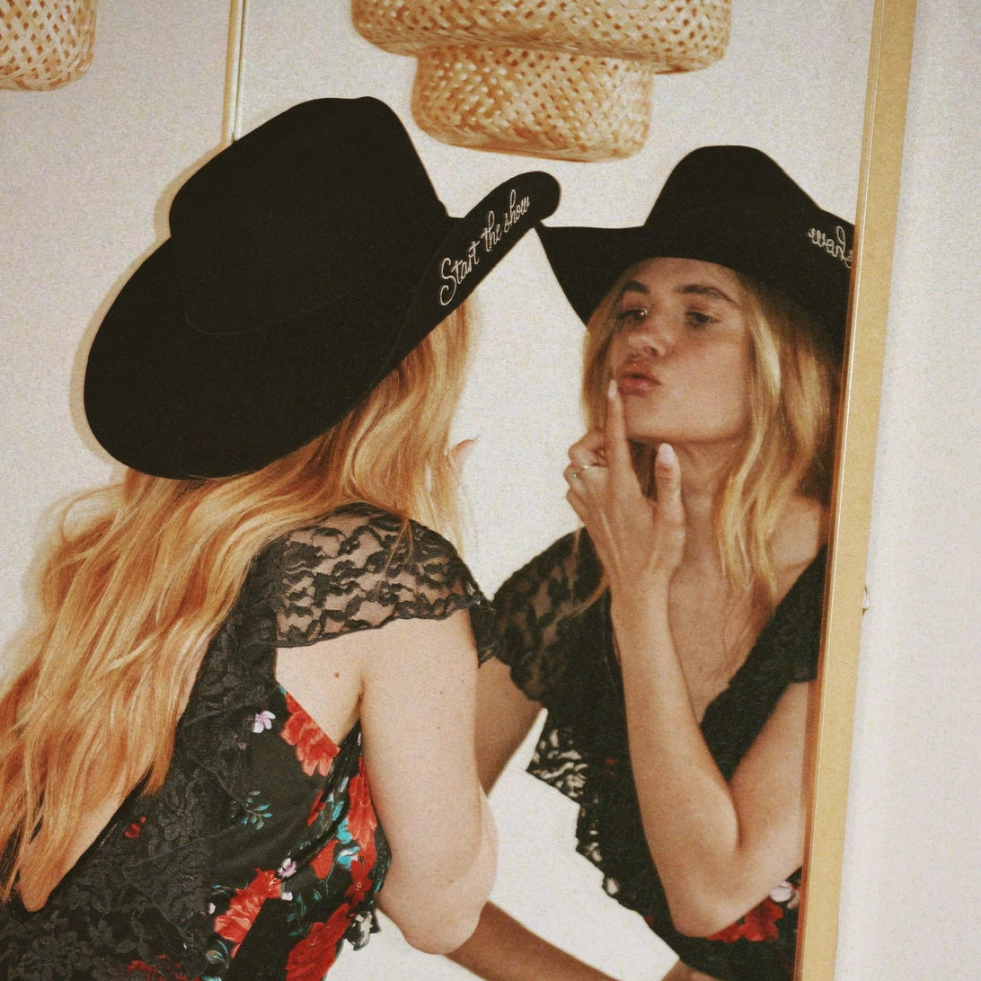 Woman wearing a black hat with text, standing in front of a mirror, looking at herself.